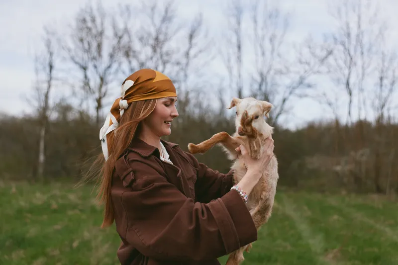 Young woman with long red hair wearing a brown coat and headscarf happily holds a playful baby goat outdoors in a grassy field during early spring.