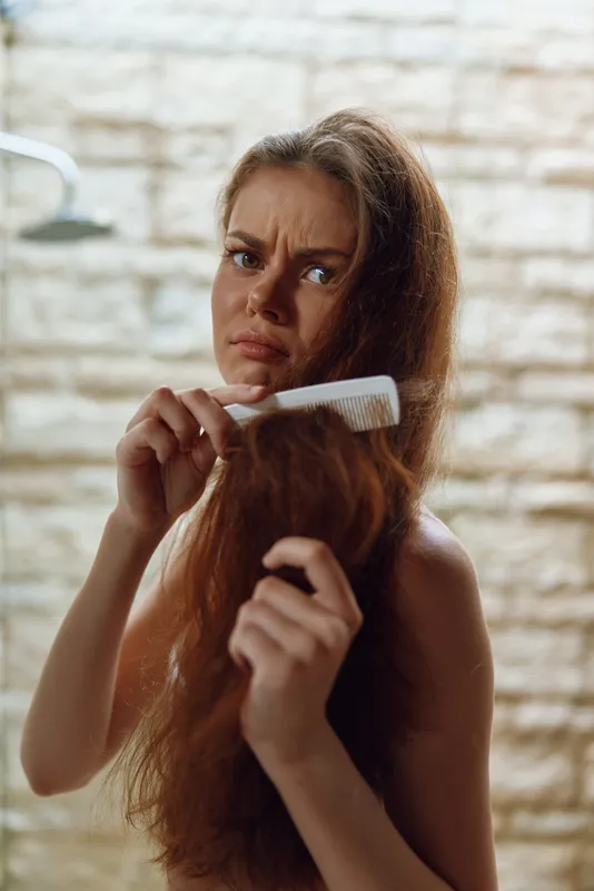 Woman with long hair struggling to comb tangled strands in bathroom with beige stone wall background, showing frustration and concentration during hair care routine.