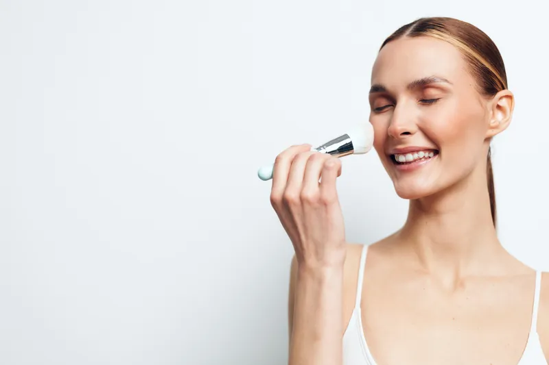 Young woman with clean skin applies makeup with a brush smiling with closed eyes in a white tank top isolated on white background, skincare and beauty routine concept.