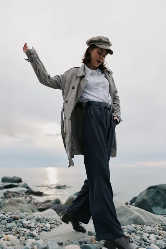 Young woman walking on rocky beach wearing casual wide pants, white shirt, textured coat, and newsboy cap. Overcast sky and calm sea create a relaxed, natural lifestyle atmosphere.