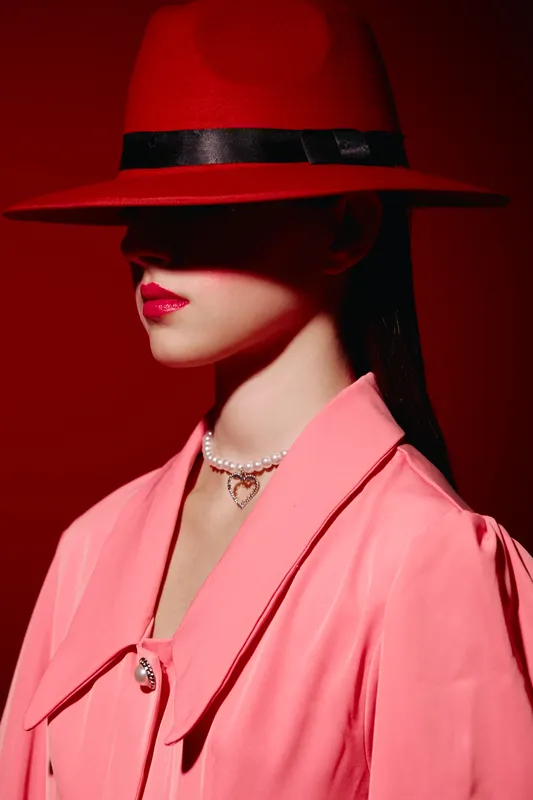 Fashionable woman wears a red hat and pink coat, with a pearl necklace, her lips painted red against a vibrant red background. Style concept with contrast and elegance.