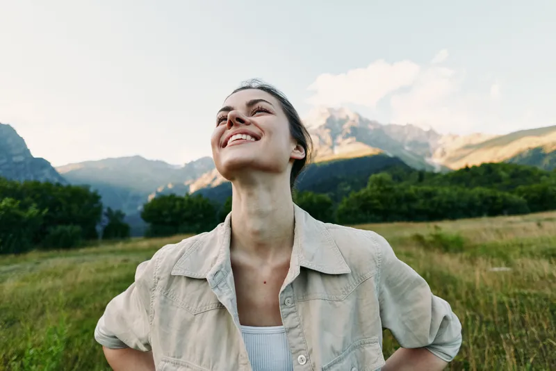 Young woman with short hair enjoys fresh mountain air with closed eyes and a smile while standing in a green meadow under a clear sky.