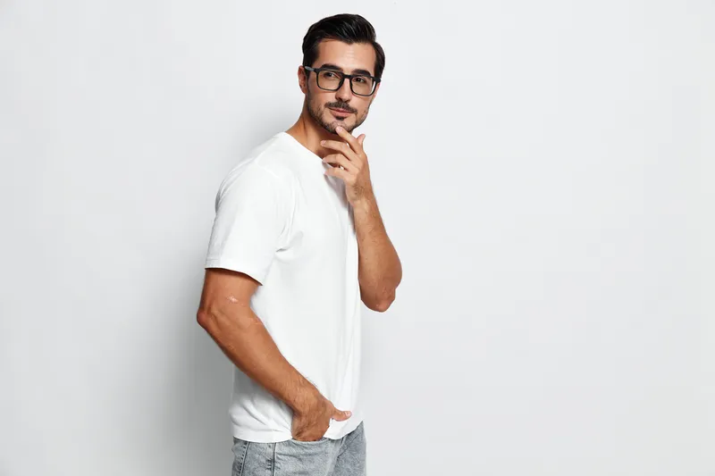 Confident young caucasian man in white t-shirt and glasses standing thoughtfully with hand on chin against a plain light background. Studio portrait concept.