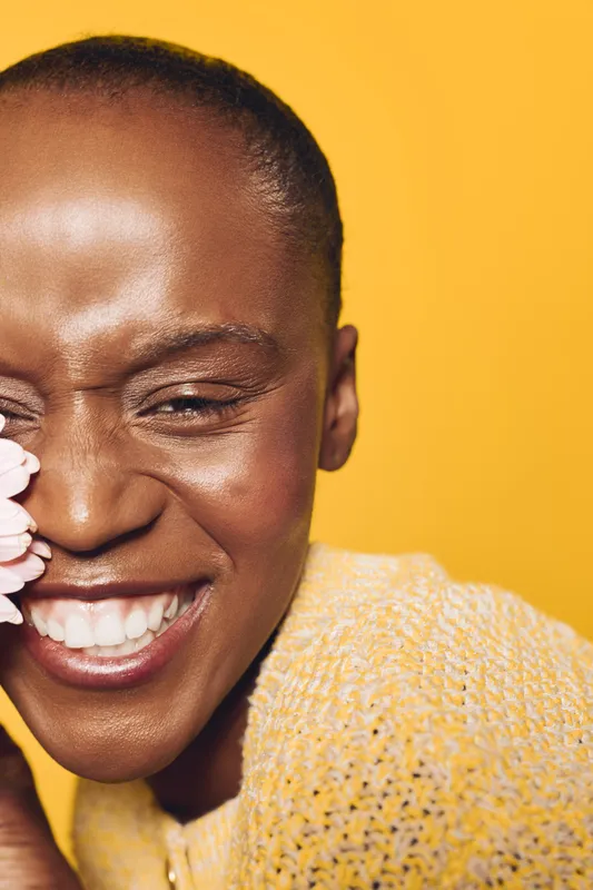 Close-up of smiling African woman with dark skin holding pink flower near face against yellow background. Concept of happiness, natural beauty, floral, and joyful emotion in studio lighting.