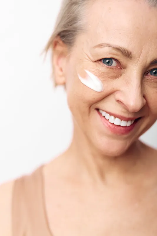 Smiling middle aged woman with blue eyes applies white face cream on cheek, close up on light background. Concept of skincare, natural beauty, anti aging, and self care routine.
