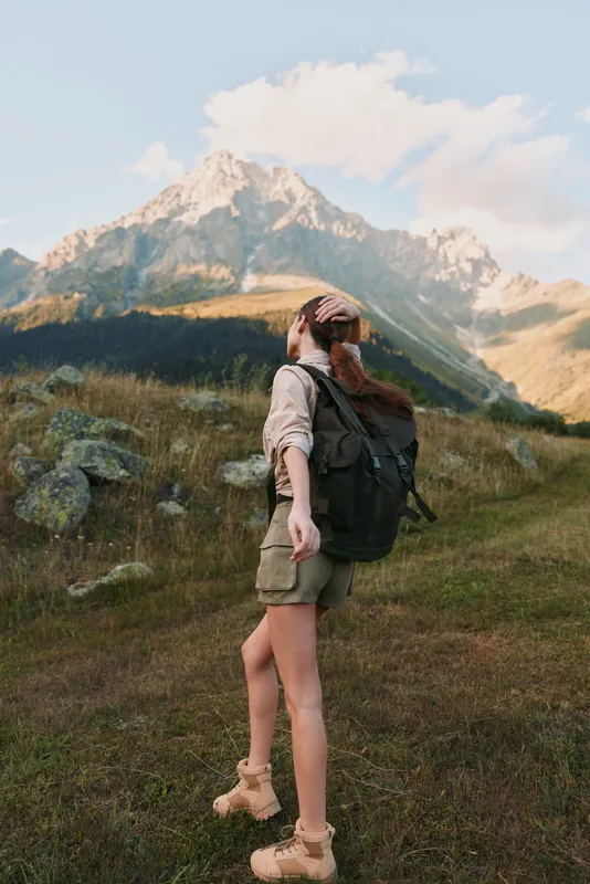 A woman hiker with a backpack stands on a grassy trail, facing distant mountains during golden hour, conveying adventure, exploration, and outdoor lifestyle.