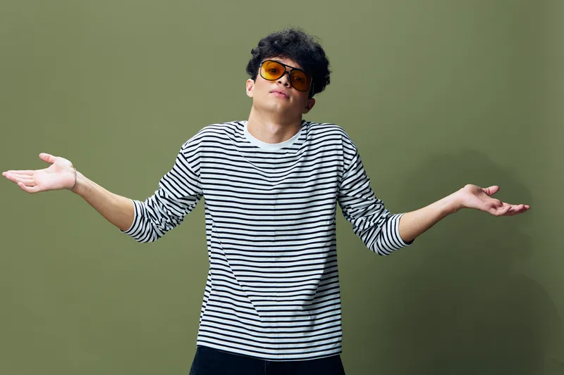 Young man in striped shirt and sunglasses shrugging with raised arms, expressing confusion and casual attitude against olive green background in soft studio light.