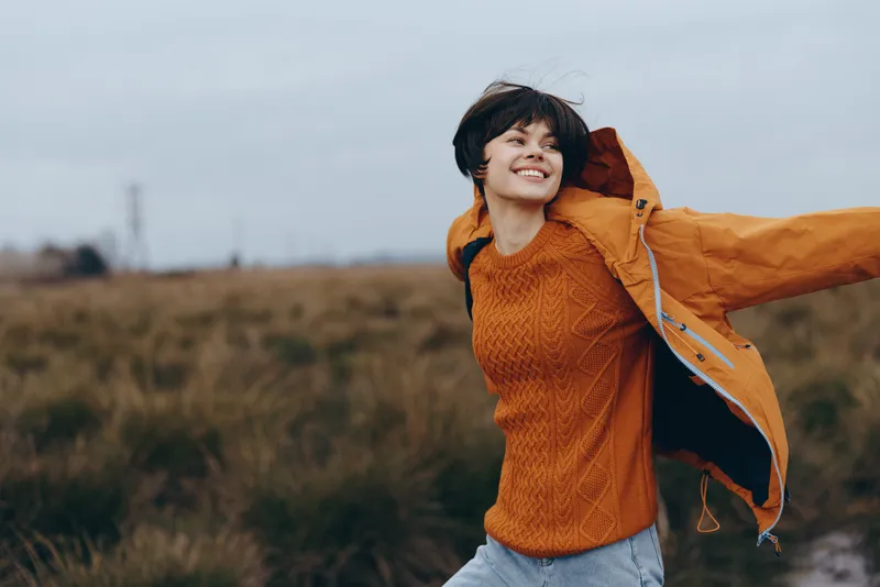Woman outdoor lifestyle, smiling in autumn field, orange jacket, facing camera, wind moves hair, cloudy sky, casual travel mood, with countryside horizon and soft natural light, today
