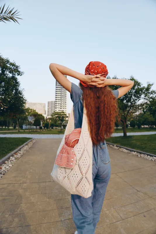 Lifestyle woman in modern boho-western denim street style with red bandana and woven bag, captured with film color quality in an urban park setting under clear sky.