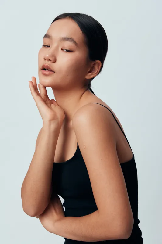 Young woman with natural skin wearing black tank top posing with closed eyes and hand near face against plain background. Beauty, fashion, minimalist style with smooth skin and serene expression.