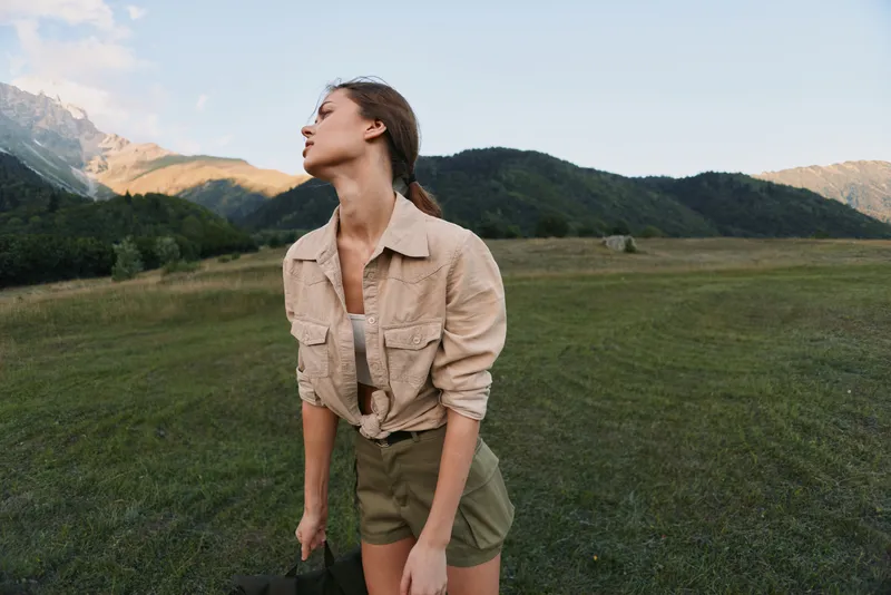 A young woman in casual clothes stands in a green field with mountains in the background, looking upwards. The scene captures outdoor lifestyle and nature exploration.