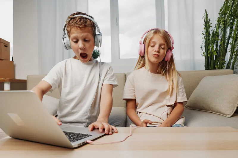 Two children with headphones are sitting on a couch in a bright room using a laptop. The boy looks surprised while the girl seems calm and focused. Concept of technology and childhood.