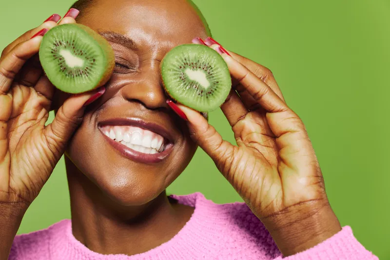 Happy dark skinned African woman with short hair holds two halves of fresh kiwi fruit near closed eyes smiling. Concept for healthy lifestyle, natural food, and joyful emotions in studio.