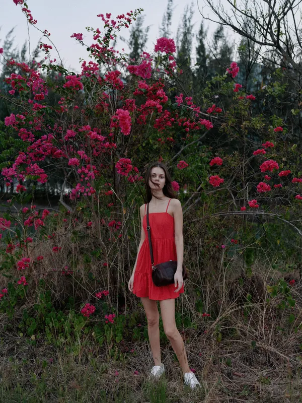 young woman in red dress standing near blooming pink flowers, natural outdoor setting with greenery and soft light, casual summer style portrait