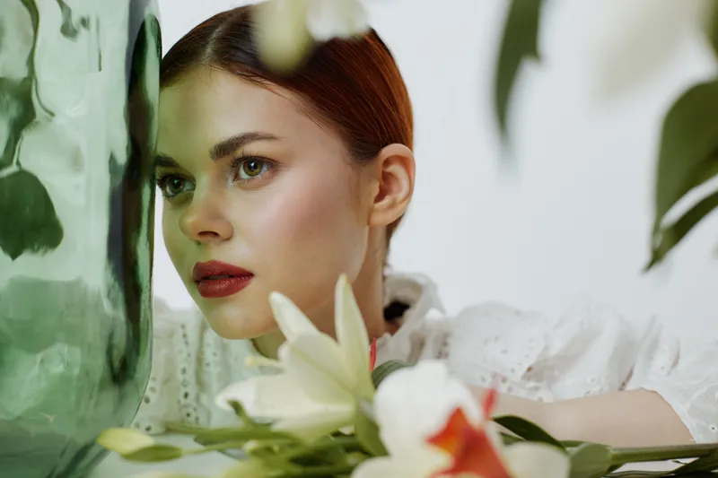 Elegant young woman with red hair posing beside a green vase and white lilies in a bright, fresh setting, embodying beauty, peace, and harmony