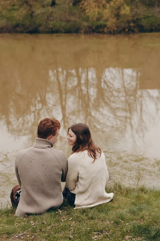 A couple sits close on a grassy bank beside a calm lake, sharing a quiet moment in sweaters. The tranquil setting highlights connection, companionship, and a cozy outdoor romance.