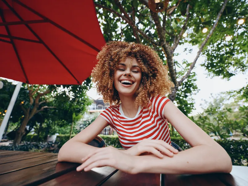 Joyful portrait of a woman with curly hair laughing at a wooden table outdoors under a red umbrella, wearing a red and white striped shirt, capturing vibrant summer energy.