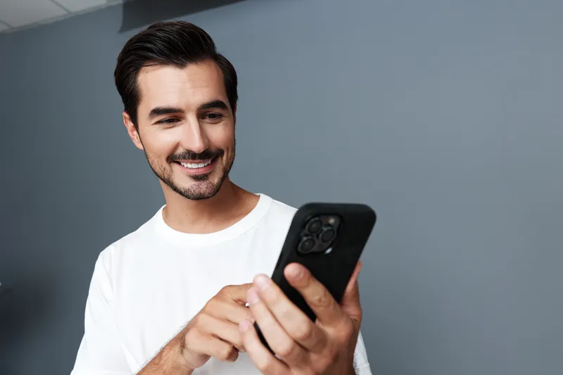 Smiling man wearing a white shirt looking at his mobile phone with a cheerful expression, standing against a solid grey background in a modern interior space.