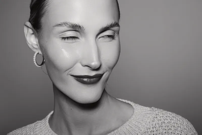 Young white woman with light skin smiles with closed eyes wearing hoop earrings and knit sweater in studio. Concept of natural beauty and positive emotions.