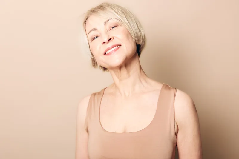 Smiling mature woman with short blonde hair wearing a beige sleeveless top poses against a light beige background. Concept of aging beauty, confidence, and natural skin care.