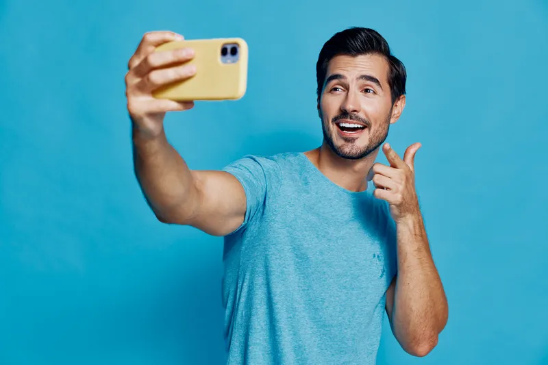 Smiling man taking a selfie with his smartphone while posing joyfully against a vibrant blue background, expressing happiness and self-confidence in casual attire.