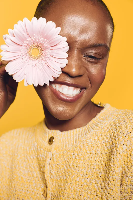 Happy African woman with dark skin smiles with closed eyes holding pink flower near face. Yellow background and yellow sweater create warm, joyful mood. Concept of beauty and nature.