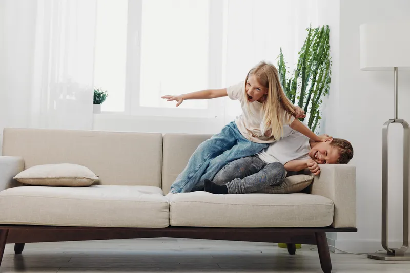 Playful children having fun on beige couch in bright living room. A girl and boy laugh while playing, showing family joy and sibling bonding at home with natural light and modern decor.