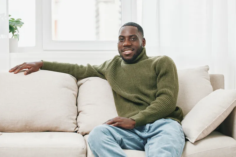 Smiling African American man in cozy green sweater relaxing on a beige sofa, representing comfort and happiness in a bright living room