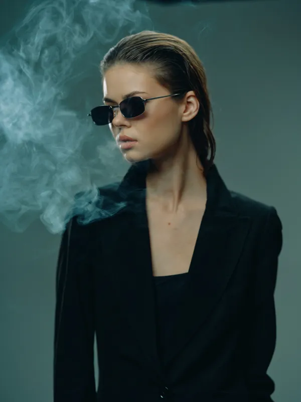 Stylish woman in black blazer wearing sunglasses with smoky background, exuding confidence and mystery.