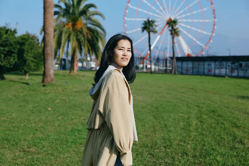 Woman wearing a beige coat in a sunny park with a ferris wheel in the background, capturing casual fashion and a cheerful mood during a leisurely urban outing