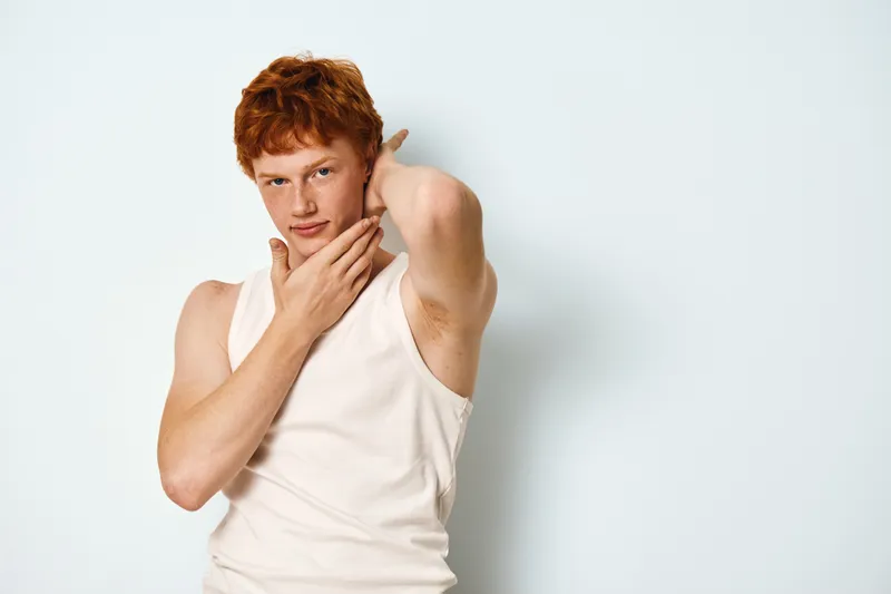 Young man with red hair wearing sleeveless shirt touching his face and neck posing thoughtful on light background. Male model, casual style with confident expression in studio lighting.
