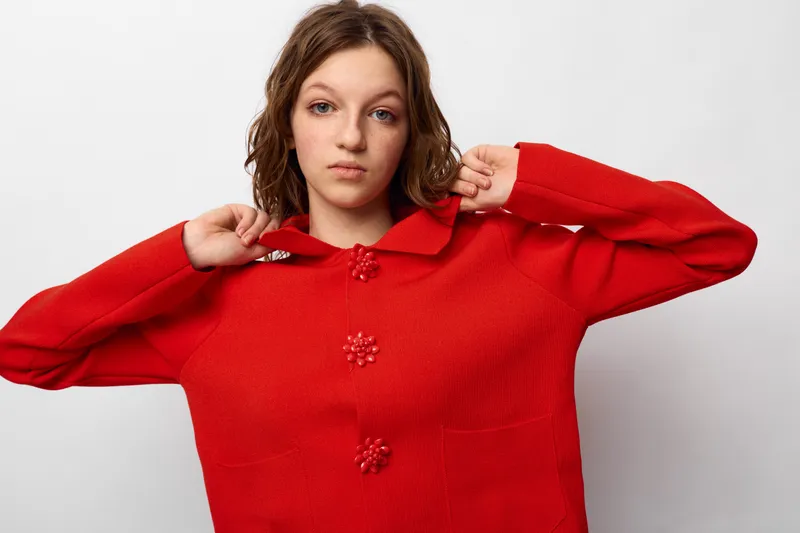 Young woman in red sweater adjusts collar while looking at camera. Casual fashion portrait with neutral expression on white background. Concept of style, clothing, and youth culture.