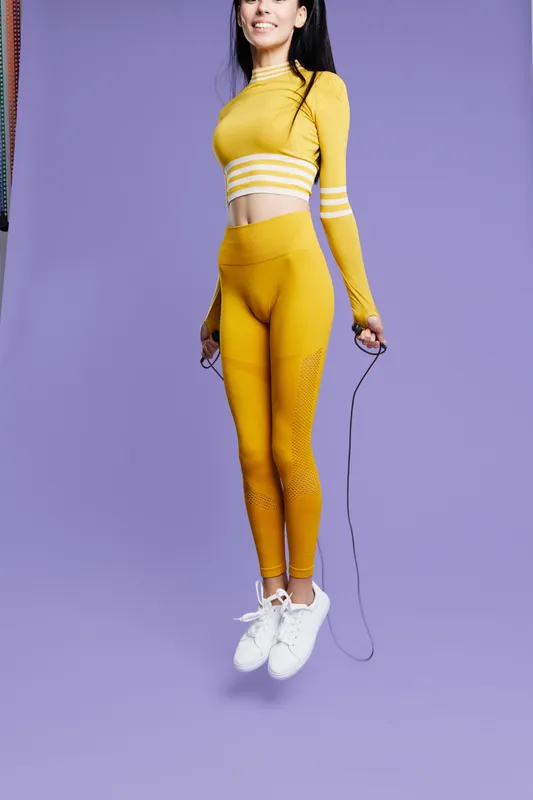 Active woman sport fitness jumping with skipping rope wearing yellow workout clothes and white sneakers on purple background, fitness motivation and healthy lifestyle concept.