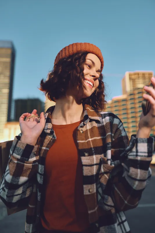 woman smiling with phone in urban city portrait wearing beanie at golden hour, candid lifestyle and authenticity captured in emotional storytelling, mindful living and warm golden hour glow