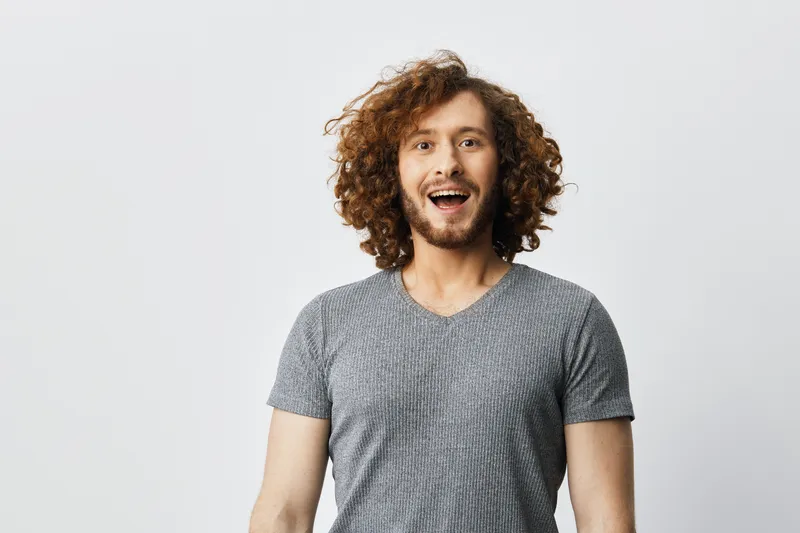 Cheerful man with curly hair wearing a gray t shirt, expressing happiness against a light background, ideal for lifestyle and personal branding concepts