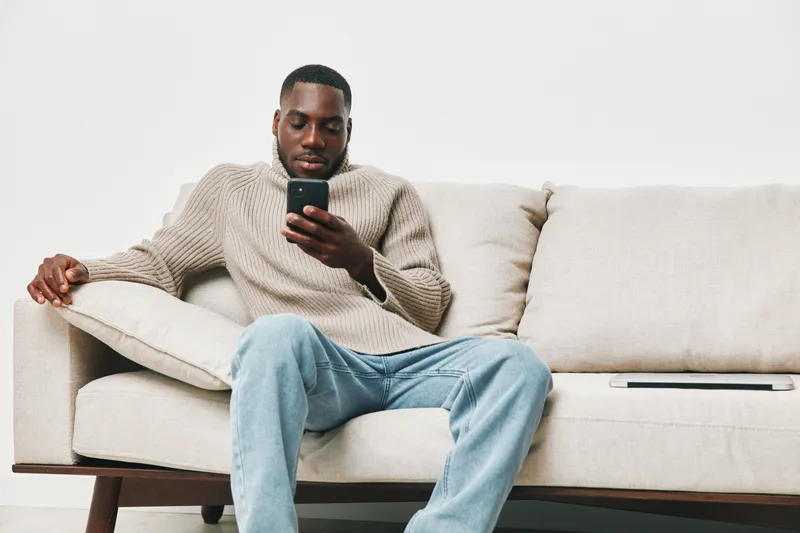 A young African American man lounging on a modern sofa, engrossed in his smartphone, wearing a cozy knit sweater and casual jeans in a minimalist living space