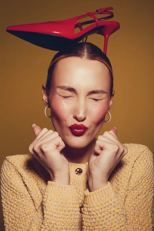 Young woman with red lipstick and closed eyes holds fists near face, balancing red high heel shoe on head. Fashion concept with playful expression and warm studio lighting.