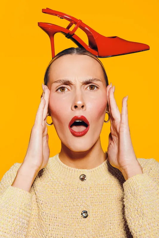 Surprised woman with red lipstick holds red high heel shoe on her head against bright yellow background. Concept of fashion, style, and unexpected moments in studio portrait.