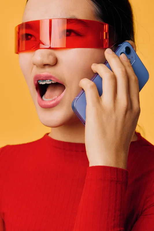 Young woman with braces wearing red futuristic glasses speaking on smartphone with open mouth expression against yellow background. Portrait, fashion and technology concept.