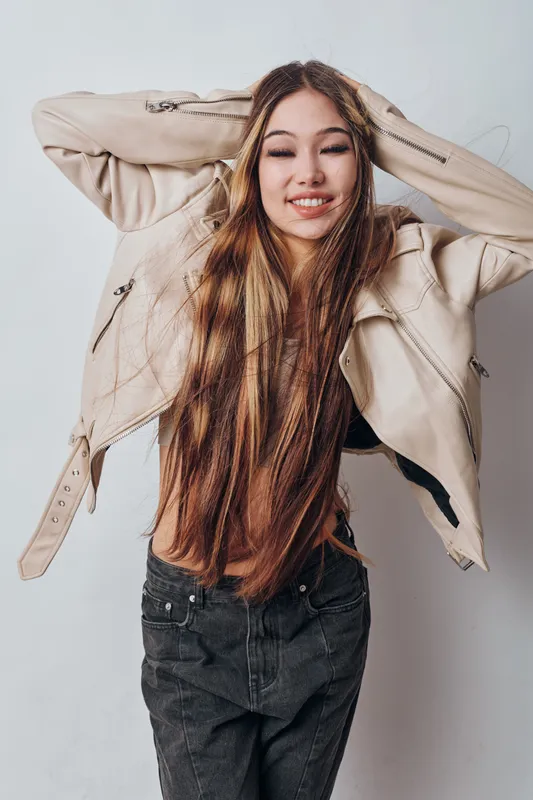 Young woman with long hair wearing beige leather jacket and black pants posing with hands behind head smiling on white background. Studio portrait of casual fashion.