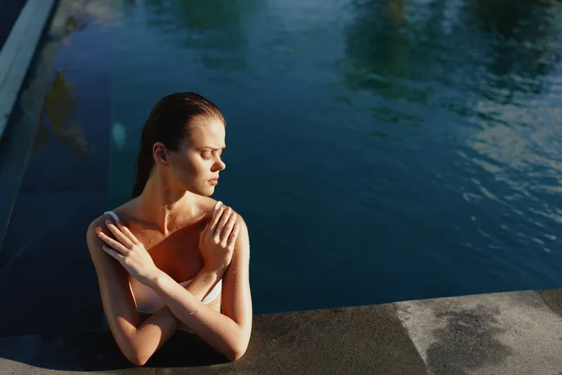 A solitary woman sits at the pool edge, arms crossed over her chest, eyes closed, absorbing the warm sun. A quiet moment of self reflection and inner calm beside sparkling blue water.