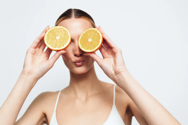 A young woman in a white top holds two orange halves in front of her eyes making a playful face on a white background. Concept for freshness, health, and fun lifestyle.