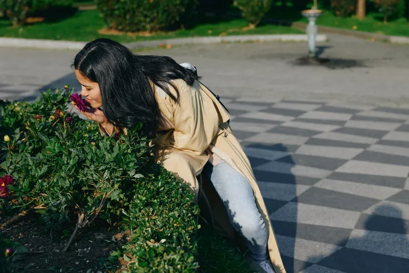Woman leans toward a flowering hedge in a park, smelling blossoms and enjoying outdoor greenery. Sunlit scene with patterned pavement and lush shrubs, casual coat, candid moment of nature.