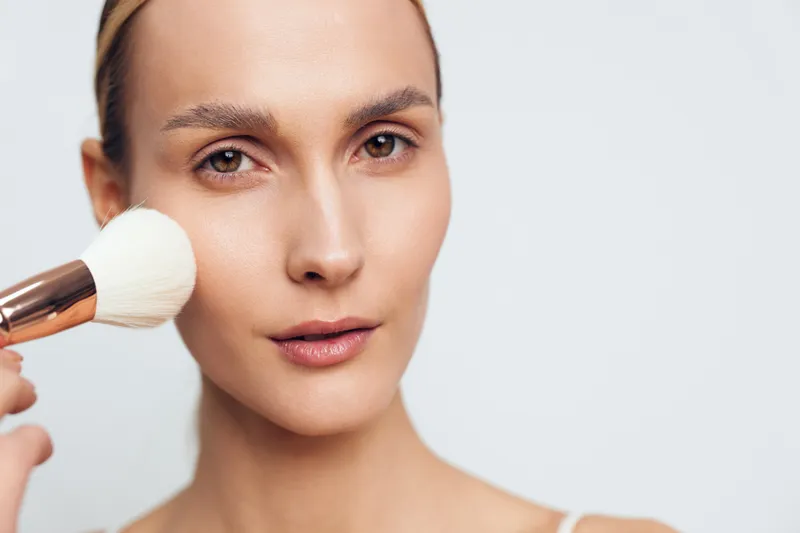 Young woman applies makeup with powder brush on clean face with natural skin tone isolated on white background, beauty routine concept with soft natural lighting and close up view.