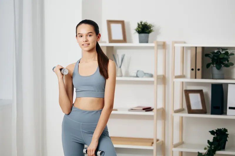Woman fitness model in a light blue athletic set stands with a dumbbell in a bright home gym, practicing strength training and balance for a focused home workout session.