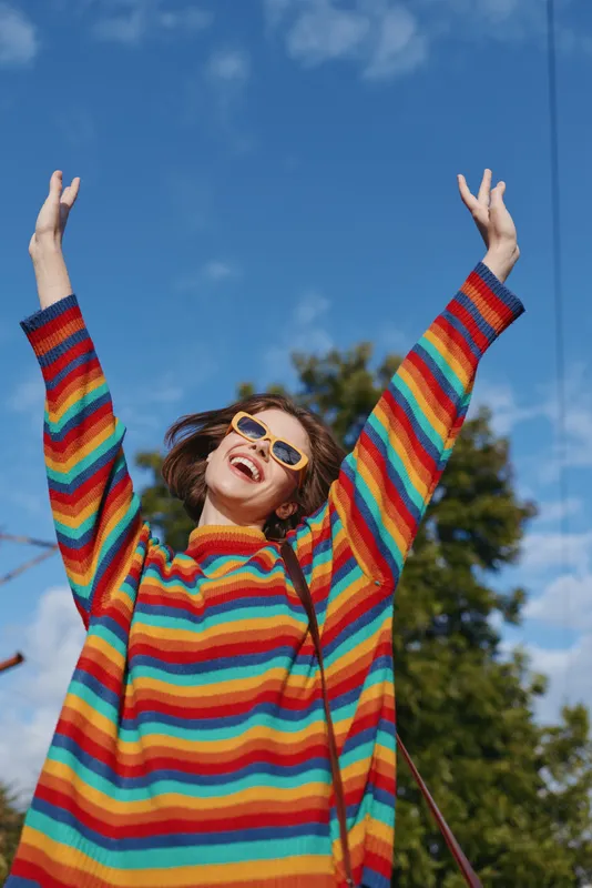 Young woman, striped sweater, arms raised, smiling, sunglasses, blue sky, outdoor, joyful — mid 20s model in colorful casual outfit celebrating under clear sky in urban park.