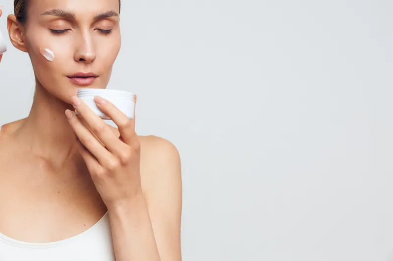 Smiling woman applying white facial cream on her cheek, close up against a light background. Concept of skincare, beauty routine, selfcare, moisturizer, wellness. Soft lighting creates a calm, fresh
