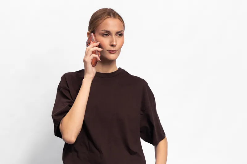 Young woman in dark t-shirt talking on smartphone, looking away with thoughtful expression. Isolated on white background. Concept of communication, technology, and casual style.