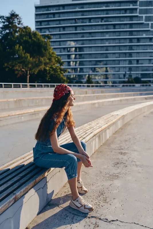 Lifestyle woman in modern boho-western denim street style sitting outdoors with sunlight, wearing a red bandana, relaxed and thoughtful, film color effect.