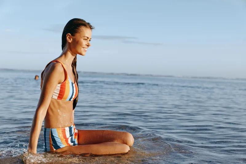Woman in striped swimsuit kneels in shallow ocean at the beach, calm morning light, practicing yoga meditation mindfulness, breathing slowly for summer wellness lifestyle concept
